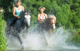 A couple of young women gleefully blast through a shallow stream while atop their trusty steeds.