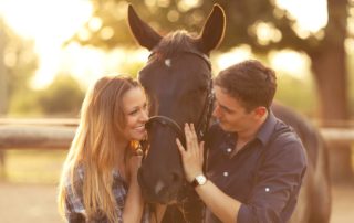 A young couple enjoys a romantic dude ranch getaway while smiling at each other with a horse in between them.