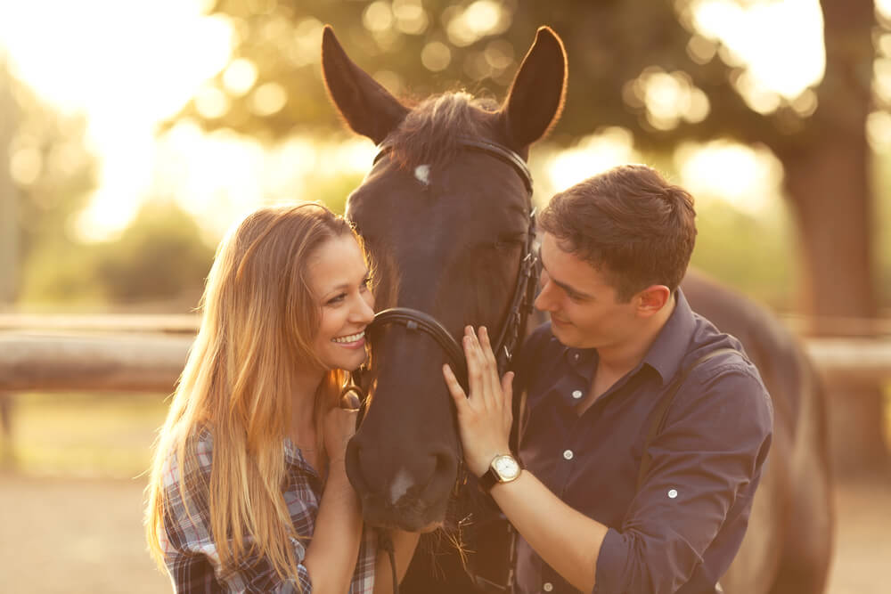 A young couple enjoys a romantic dude ranch getaway while smiling at each other with a horse in between them.